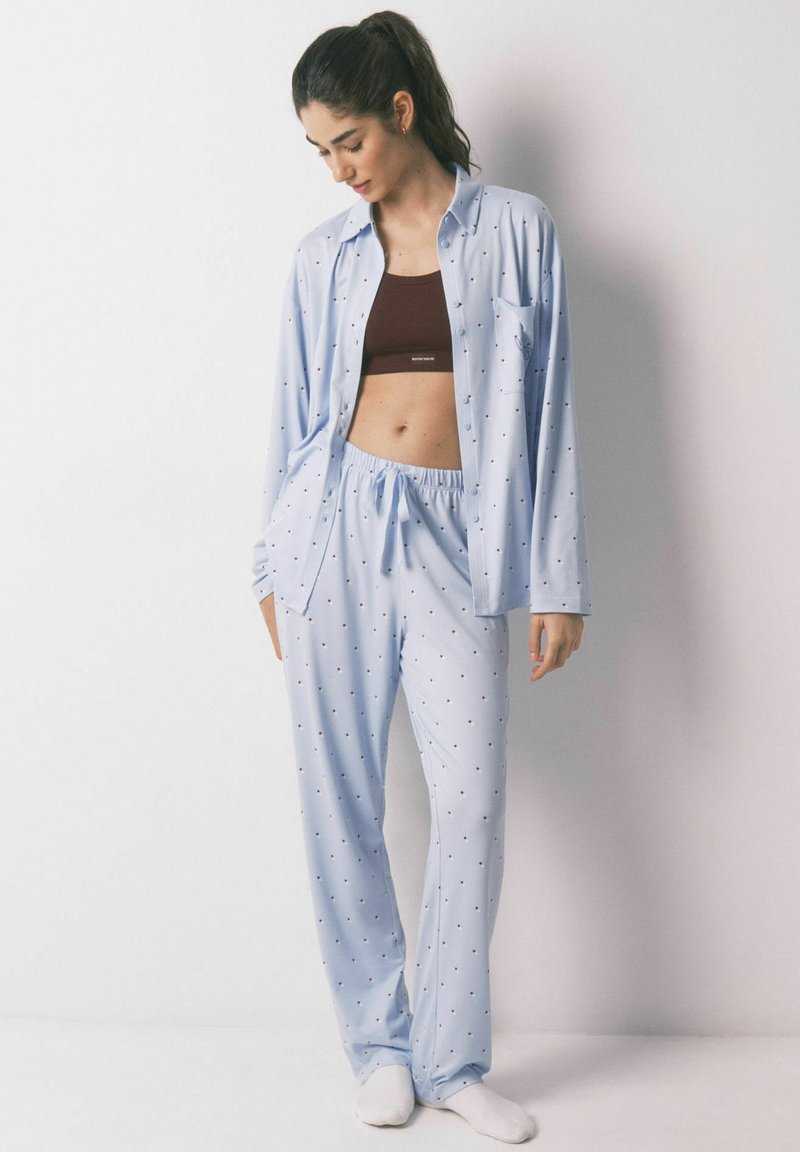 Femme debout contre un mur, portant un pyjama bleu clair à pois par-dessus un crop top marron et des chaussettes blanches, regardant vers le bas.