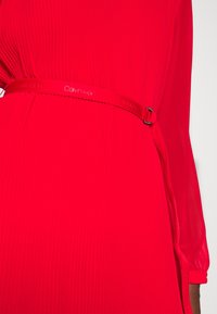 Punainen mekko, jossa on ribbikuvio, pitkät hihat ja kiiltävä, merkitty vyö. Suunnittelu korostaa istuvaa siluettia.