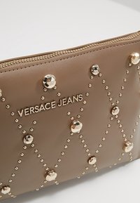 Pochette marron Versace Jeans avec des clous dorés disposés en motifs en losange et une fermeture éclair dorée sur le bord supérieur.