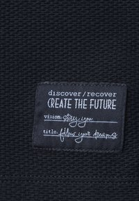 Mustan tekstuurin kangas, jossa on ommeltu etiketti, jossa on valkoista tekstiä. Etiketissä on lauseet "discover/recover", "CREATE THE FUTURE" ja inspiraatiota tarjoavia otsikoita.