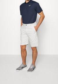 Marinblå polotröja med krage och Nike-logotyp, kombinerad med vita shorts som har framfickor. Grå sportskor med svarta detaljer.