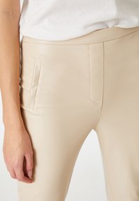 Beige, tætsiddende bukser lavet af glat, strækbart materiale. Har en sidelomme, minimal syning og et rent, strømlinet design.
