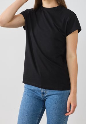 Frau trägt ein schlichtes schwarzes Kurzarm-T-Shirt und blaue Jeans, mit einem Arm nahe am Kopf erhoben und dem anderen seitlich ruhend.