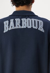 Sötétkék póló, hátul fehér hímzett "BARBOUR" felirattal, klasszikus gallérral és sima, texturált anyagból készült.