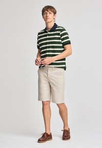 Grön och vitrandig polo med marinblå krage; ljusbeige shorts; bruna läderskor. Modellen står med händerna sammanflätade, neutral bakgrund.