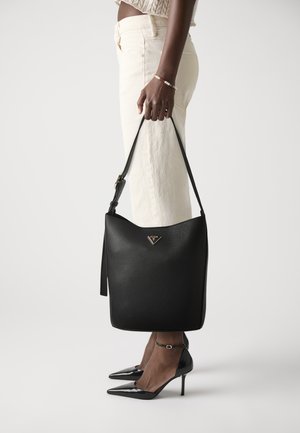 Zwarte leren tote bag met een gestructureerde afwerking en gouden logo-detail, vastgehouden door een persoon in crèmekleurige broek en zwarte puntige hakken.
