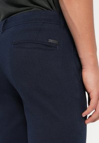 Pantaloni in tessuto a spina di pesce blu navy con tasca posteriore e etichetta del marchio. Texture liscia con vestibilità sartoriale e cuciture definite.