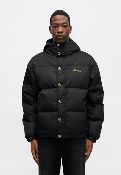 Melnā pūkainā jaka ar kapuci, ar zeltainiem poga un mazu zeltainu Versace logotipu kreisajā krūšu daļā. Tekstūras, iešūta dizaina.