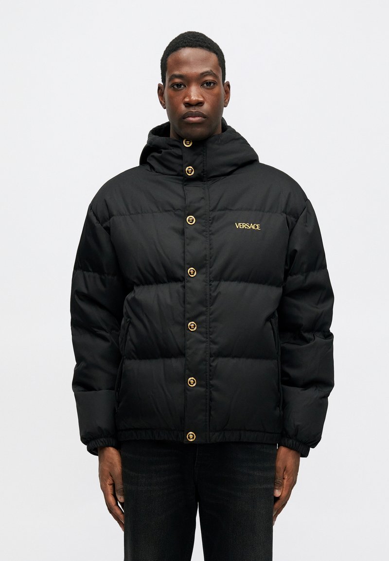 Sort pufferjakke med hætte, der har guldhardwareknapper og et lille guld Versace-logo på venstre bryst. Takket, quiltet design.