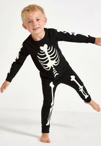 Čierny súbor pyžama s dlhým rukávom, ktorý má na prednej strane a na nohaviciach bielu potlač kostry. Jemná tkanina, priliehavý strih s rebrovanými manžetami.