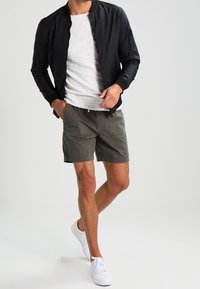 Svart bomberjacka, grå t-shirt och olivgröna shorts. Modell som bär vita sneakers. Släta material och avslappnad design.