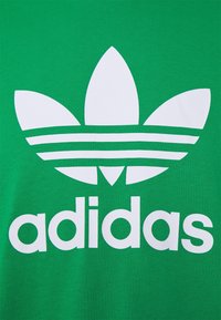 Grön tyg med stor vit Adidas trefoil-logotyp och texten "adidas" tryckt under logotypen.