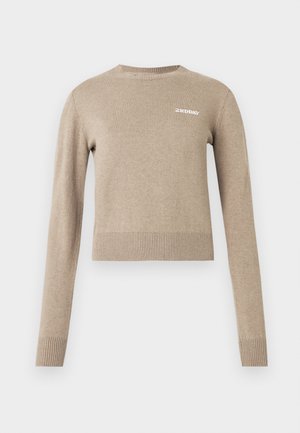 Pull tricoté beige à manches longues, col rond, poignets et ourlet côtelés, avec un petit logo texte blanc sur le devant.