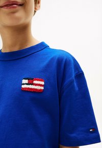 Blaues Baumwoll-T-Shirt mit einem strukturierten Patch eines Flaggen Designs in Rot, Weiß und Schwarz auf der Brust und einem kleinen Logo auf dem Ärmel.