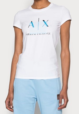Biała koszulka slim fit z krótkim rękawem i niebieskim logo A|X Armani Exchange, noszona przez osobę i zestawiona z jasnoniebieskimi spodniami.