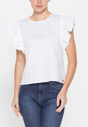 Donna con indosso una camicetta bianca con maniche corte arricciate e jeans blu in denim, in piedi su uno sfondo bianco semplice.