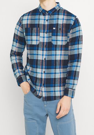Mann trägt ein blau-grau kariertes Hemd mit Knöpfen und zwei Brusttaschen sowie hellblaue Hose, steht mit einer Hand in der Hosentasche.