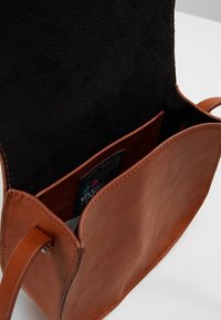 Borsa a tracolla in pelle marrone con patta aperta che mostra la fodera interna nera e un'etichetta logo visibile nella tasca interna.