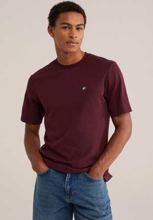 Joven con camiseta de color rojo oscuro con un pequeño logo de pájaro y vaqueros azules, de pie con las manos en los bolsillos frente a un fondo liso.