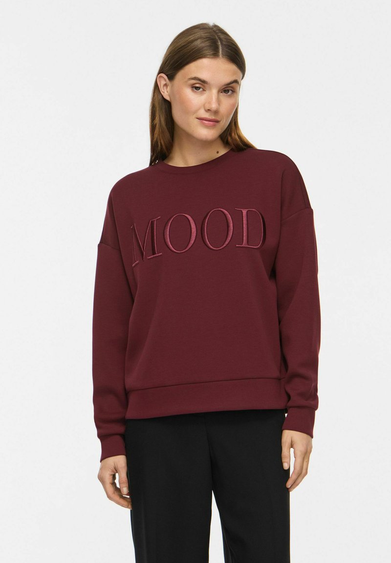 Jeune femme aux cheveux bruns portant un sweat-shirt bordeaux avec le mot "MOOD" brodé sur la poitrine et un pantalon noir.