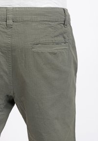 Närbild av en person som bär gröna, texturerade byxor med en horisontell slitsficka bak och bältesöglor på midjan.