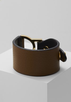Bracelet manchette en cuir marron avec large bande et fermoir en métal doré, présenté sur un socle blanc sur fond gris.