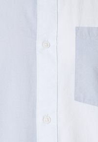 Camicia button-up azzurro chiaro e bianca con una texture liscia, caratterizzata da una tasca sul petto di un tono leggermente più scuro e bottoni bianchi.