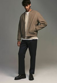 Veste bomber marron en tissu tissé, portée sur un pull beige et un pantalon noir. Chaussures noires à lacets avec une semelle épaisse. Design épuré et minimaliste.