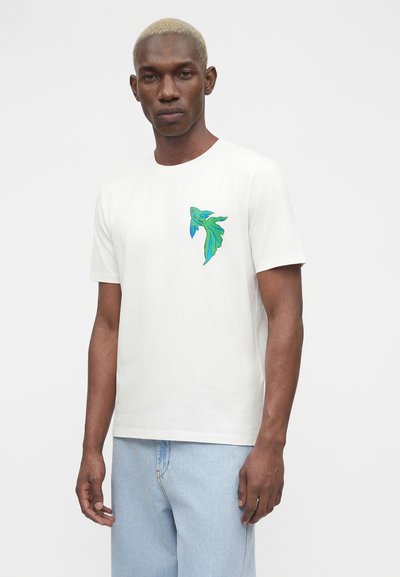 Άνδρας με κοντά ξανθά μαλλιά που φοράει λευκό μπλουζάκι με πράσινο και μπλε πουλί στο στήθος και ανοιχτό μπλε τζιν μπροστά σε απλό φόντο.