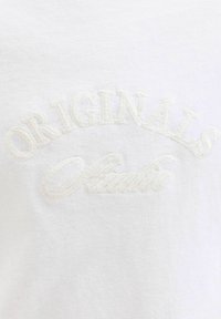 Camiseta blanca de algodón con texto bordado que dice "ORIGINALS Studio" en un hilo sutil y claro. Textura suave, diseño sencillo.