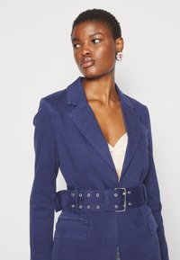 Ναυτικό μπλέ βαμβακερό σακάκι με ζώνη στη μέση, που διαθέτει κολάρο με αυλάκι, δύο μπροστινές τσέπες και απαλό ύφασμα.