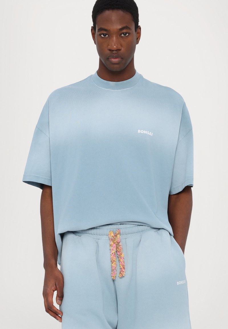 Helles blaues Oversized-T-Shirt mit rundem Ausschnitt und kurzen Ärmeln. Verziert mit dem weißen "BONSAI"-Logo. Kombiniert mit passenden Jogginghosen und buntem Kordelzug.