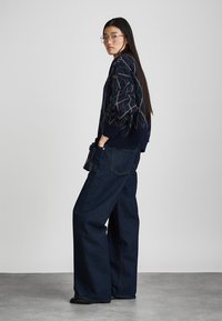 Σακάκι με ναυτικό μοτίβο και διαμαντένιο σχέδιο, συνδυασμένο με φαρδιά τζιν παντελόνια σκούρου χρώματος. Το μοντέλο κρατά μια μαύρη τσάντα και στέκεται μπροστά από ουδέτερο φόντο.