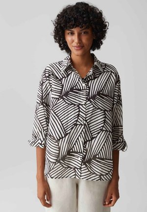 Vrouw met krullend haar, die een zwart-wit geometrisch patroon overhemd met knopen en een lichte broek draagt, staand tegen een effen achtergrond.