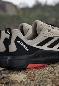 Beżowe buty sportowe z czarnymi akcentami, teksturowana cholewka, wzór z trzema paskami i czerwoną podeszwą. Posiadają solidną konstrukcję odpowiednią do użytku na zewnątrz.