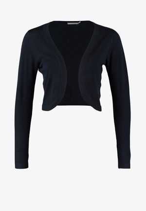 Femme portant une robe beige longueur genoux avec un cardigan noir court, des collants noirs transparents, des talons noirs, et tenant une pochette noire.