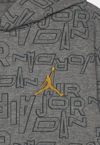Grauer Hoodie mit einem strukturierten Muster des Wortes "JORDAN" in Dunkelgrau. Verfügt über ein gelbes, gesticktes Jumpman-Logo auf der linken Brust.