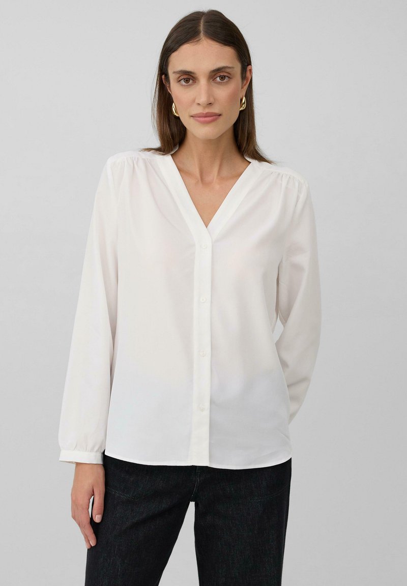 Blusa bianca a maniche lunghe con scollo a V, dotata di una chiusura con bottoni sul davanti, in tessuto morbido e con dettagli arricciati sulle spalle.