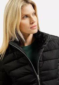 Czarna kurtka puchowa z pikowanymi sekcjami i srebrnym zamkiem. Miękka, czarna futrzana podszewka przy kołnierzu. Noszona na zielonej bluzce.