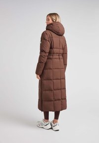 Abrigo de plumas largo marrón con capucha, presenta un diseño acolchado y cremalleras laterales. Lleva puesto sobre leggings oscuros y zapatillas blancas.