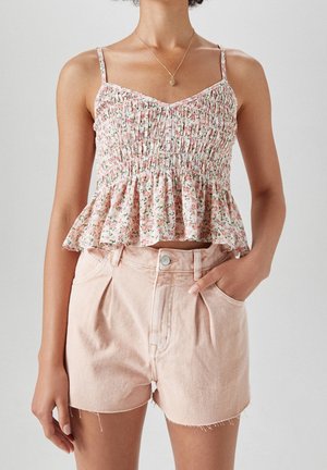 Vrouw die een mouwloze, bloemrijke, gesmokte crop top draagt en lichtroze, hooggetailleerde denim shorts, met één hand in de zak en een ketting om.