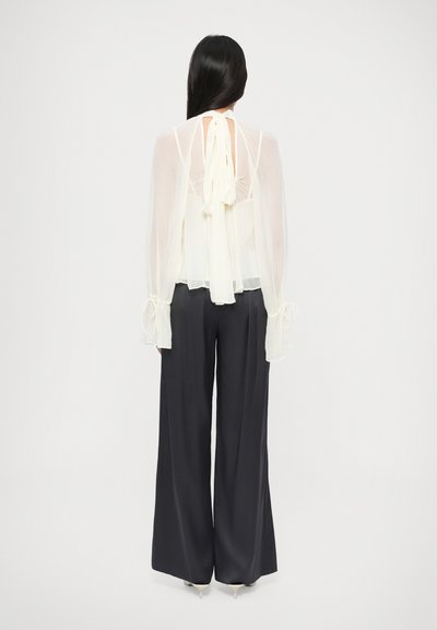 Kerge läbipaistev kreemjas pluus, millel on tagant seotud detail, kombineeritud tumedate laia säärega pükstega. Pluusil on õhulised varrukad lipsudetailidega.