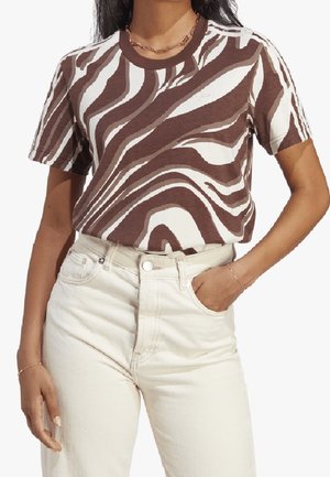 Kobieta ubrana w t-shirt w brązowo-biały abstrakcyjny wzór, włożony do wysokich, kremowych jeansów, z jedną ręką w kieszeni.