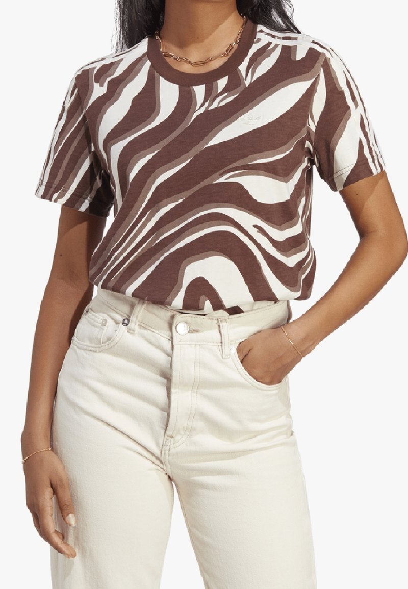 Mujer con camiseta de patrón abstracto marrón y blanco metida en vaqueros de cintura alta color crema, con una mano en el bolsillo.