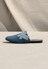 Μπλε πάνινο παπούτσι τύπου slip-on με γαλάζιο φιόγκο και μικρή χρυσή καρδιά, παρουσιάζεται σε ουδέτερο φόντο με υφασμάτινο υπόβαθρο.