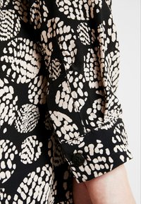 Chemise noire à manches longues avec des motifs de cercles irréguliers beiges et un bouton noir sur le poignet, portée sur le bras d'une personne sur fond blanc.