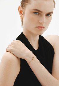 Pulsera de oro con charm de luna creciente, cadena delicada, modelo vistiendo una blusa sin mangas negra, mano descansando en el hombro. Textura suave.