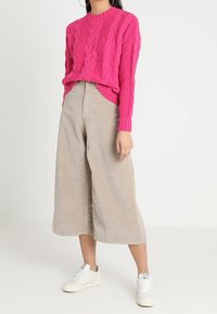 Femme portant un pull en maille torsadée rose vif, un pantalon large beige raccourci et des baskets blanches sur un fond clair uni.