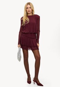 Robe bordeaux à pois avec manches longues en voile transparent, ourlet à volants superposés, associée à des escarpins rouges à talons aiguilles et une pochette argentée.