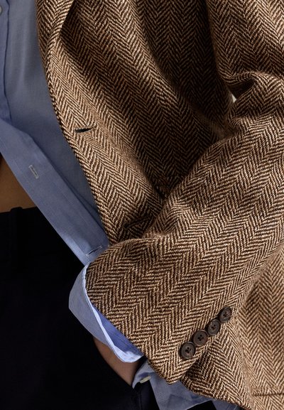 Brown herringbone patterned blazer sleeve with four dark buttons worn over a light blue shirt cuff and dark pants.